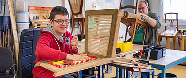 In den Caldener Werkstätten der Baunataler Diakonie Kassel hauchen Mitarbeitende alten Stühlen neues Leben ein. Unser Foto zeigt Julian Blumenstein bei der Arbeit. Im Hintergrund ist Thomas Backhaus beim Flechten zu sehen. (Foto: medio.tv/Schauderna)
