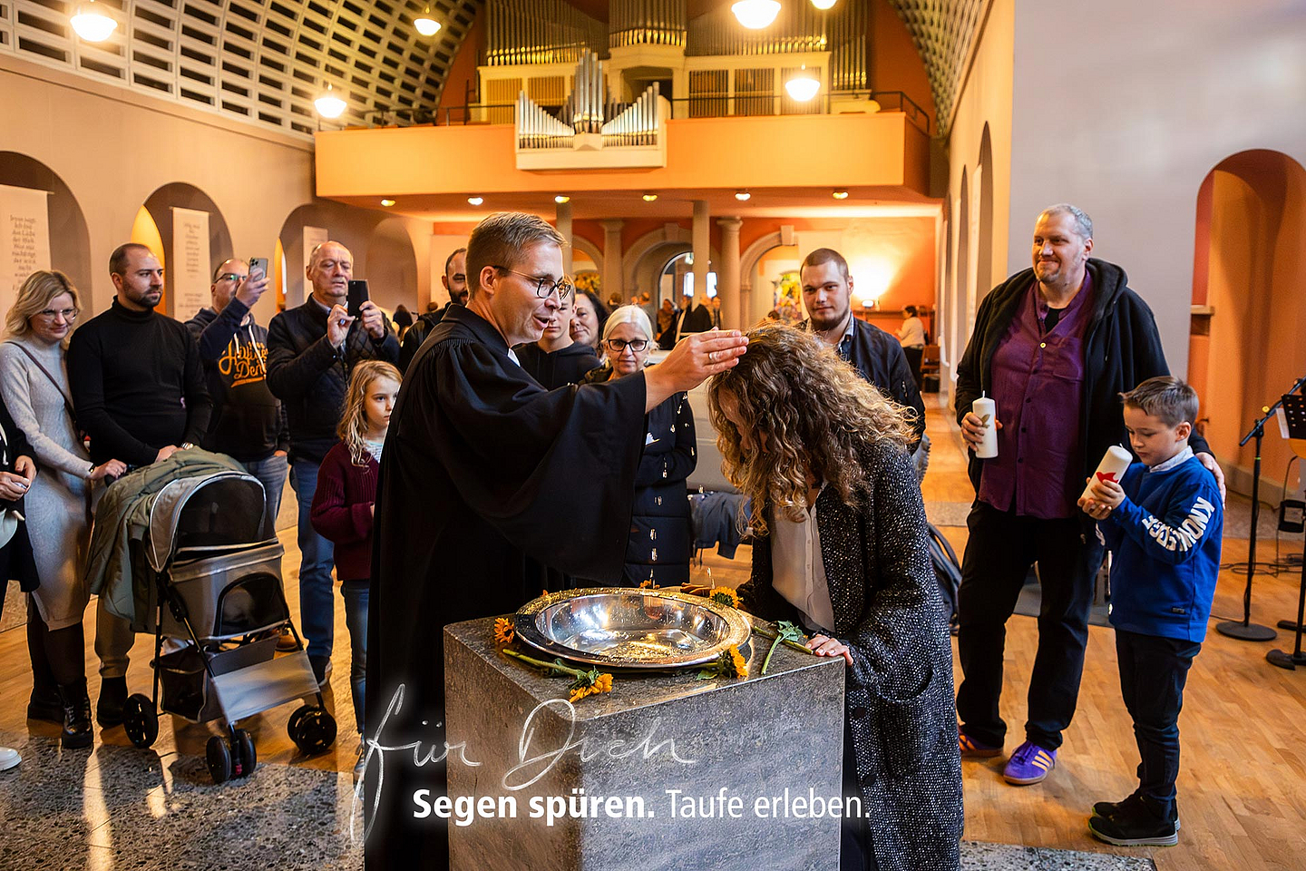 Eine Frau wird in einer evangelischen Kirche am Taufbecken von einem Pfarrer mit Wasser getauft. Um sie herum stehen Erwachsene und Kinder. Im Hintergrund sind Kirchenbänke, eine Orgel und ein Kinderwagen zu sehen. Text im Bild: ‚Für Dich. Segen spüren. Taufe erleben.