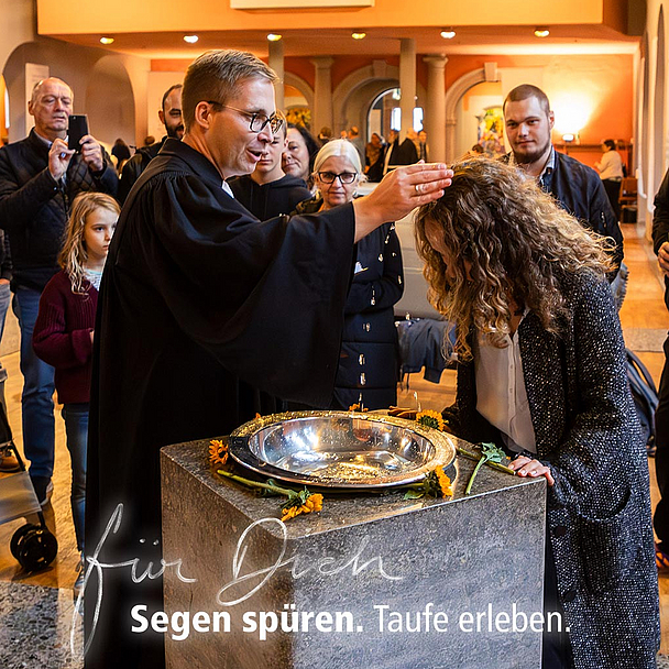 Eine Frau wird in einer evangelischen Kirche am Taufbecken von einem Pfarrer mit Wasser getauft. Um sie herum stehen Erwachsene und Kinder. Im Hintergrund sind Kirchenbänke, eine Orgel und ein Kinderwagen zu sehen. Text im Bild: ‚Für Dich. Segen spüren. Taufe erleben.