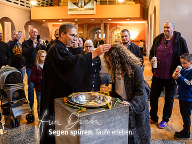 Eine Frau wird in einer evangelischen Kirche am Taufbecken von einem Pfarrer mit Wasser getauft. Um sie herum stehen Erwachsene und Kinder. Im Hintergrund sind Kirchenbänke, eine Orgel und ein Kinderwagen zu sehen. Text im Bild: ‚Für Dich. Segen spüren. Taufe erleben.