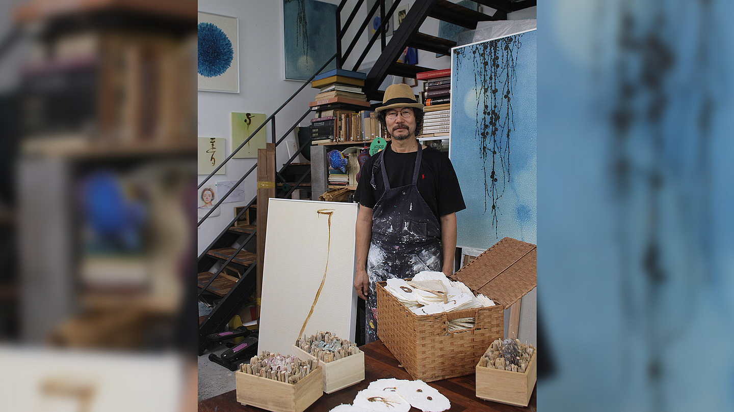 Das Foto zeigt den Künstler Woo-Hyun Lee in einem Atelier. Er steht vor einem Tisch, auf dem mehrere Körbe mit Kunstmaterialien und ein geöffneter Korb mit Papierstücken liegen. Im Hintergrund befinden sich Regale mit Büchern und Kunstwerken. Eine Treppe führt nach oben, und an der Wand hängen verschiedene Gemälde. Woo-Hyun Lee trägt eine Schürze über seiner Kleidung.