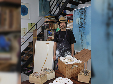 Das Foto zeigt den Künstler Woo-Hyun Lee in einem Atelier. Er steht vor einem Tisch, auf dem mehrere Körbe mit Kunstmaterialien und ein geöffneter Korb mit Papierstücken liegen. Im Hintergrund befinden sich Regale mit Büchern und Kunstwerken. Eine Treppe führt nach oben, und an der Wand hängen verschiedene Gemälde. Woo-Hyun Lee trägt eine Schürze über seiner Kleidung.