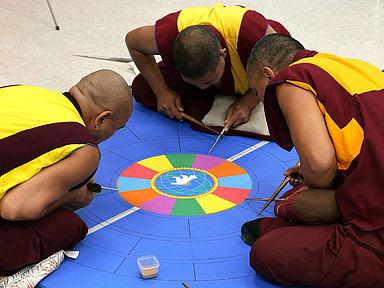 Drei Personen in traditionellen Gewändern sitzen auf dem Boden und gestalten ein farbenfrohes Sandmandala. In der Mitte des Mandalas ist ein weißer Vogel zu sehen, umgeben von bunten Segmenten. Die Personen arbeiten mit speziellen Werkzeugen an der feinen Struktur des Mandalas.