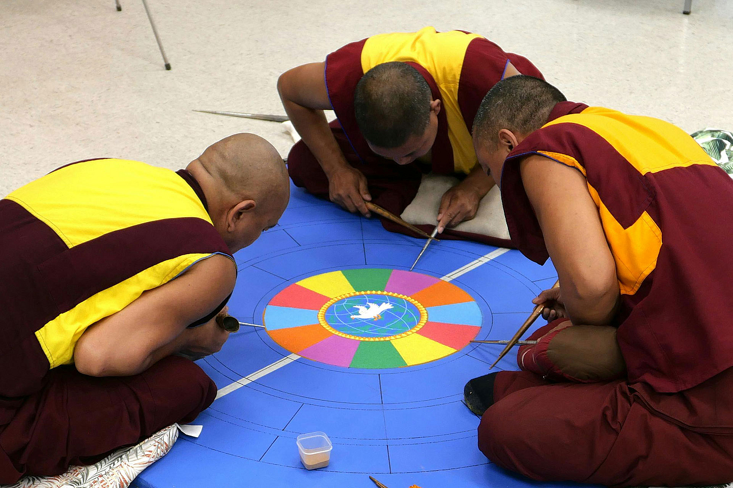 Drei Personen in traditionellen Gewändern sitzen auf dem Boden und gestalten ein farbenfrohes Sandmandala. In der Mitte des Mandalas ist ein weißer Vogel zu sehen, umgeben von bunten Segmenten. Die Personen arbeiten mit speziellen Werkzeugen an der feinen Struktur des Mandalas.