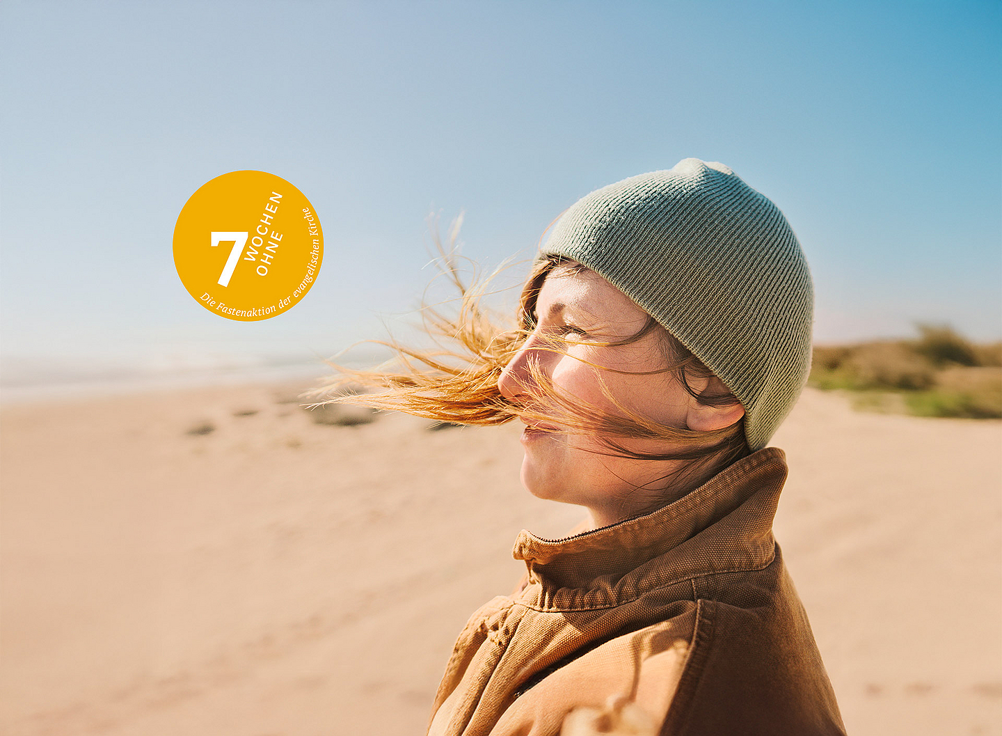 Das Motiv der Fastenaktion 2025 zeigt eine Frau, die in warmer Kleidung im Wind an einem Strand steht.