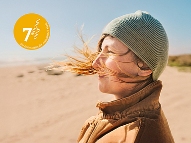 Das Motiv der Fastenaktion 2025 zeigt eine Frau, die in warmer Kleidung im Wind an einem Strand steht.