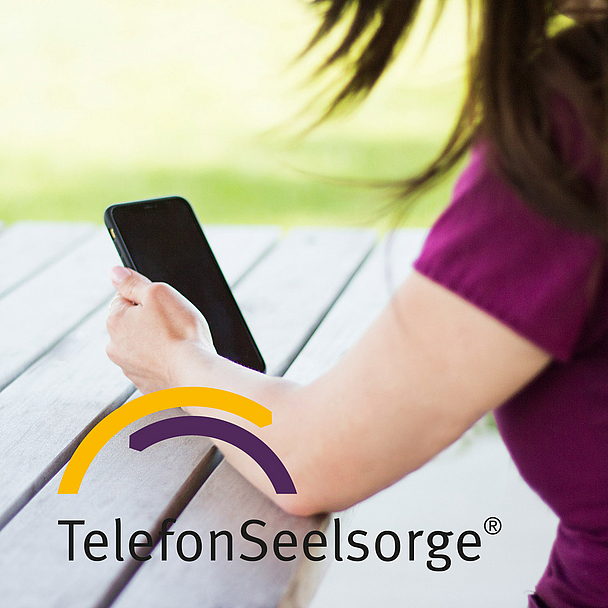 Eine Person sitzt an einem Holztisch im Freien und hält ein Smartphone in der Hand. Im Bild ist das Logo der TelefonSeelsorge zu sehen. Das Foto steht symbolisch für die telefonische und digitale Beratung von Menschen in Krisen.