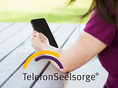 Eine Person sitzt an einem Holztisch im Freien und hält ein Smartphone in der Hand. Im Bild ist das Logo der TelefonSeelsorge zu sehen. Das Foto steht symbolisch für die telefonische und digitale Beratung von Menschen in Krisen.