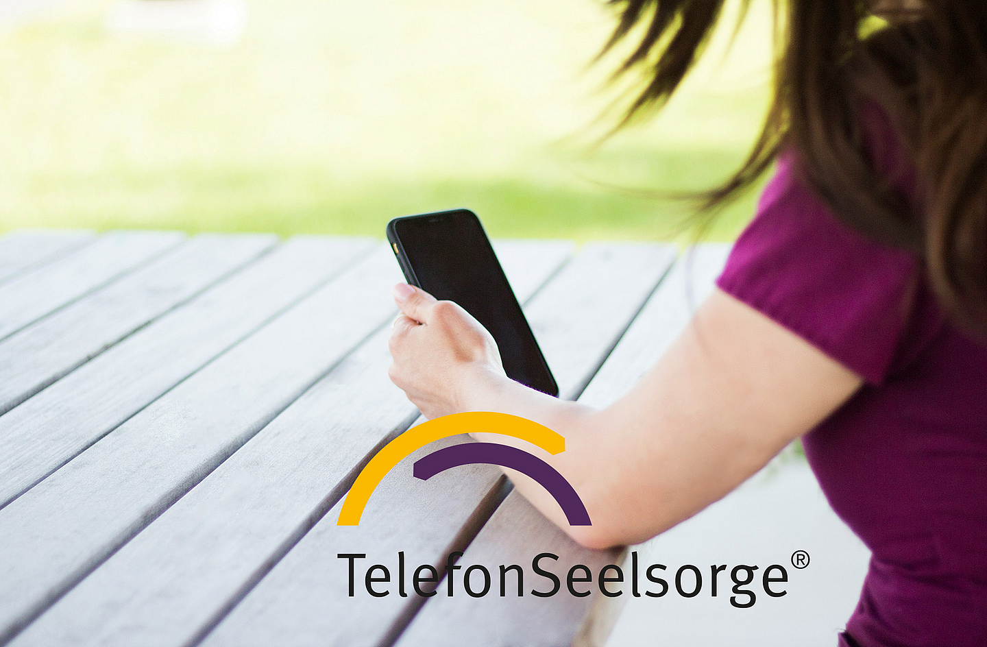 Eine Person sitzt an einem Holztisch im Freien und hält ein Smartphone in der Hand. Im Bild ist das Logo der TelefonSeelsorge zu sehen. Das Foto steht symbolisch für die telefonische und digitale Beratung von Menschen in Krisen.