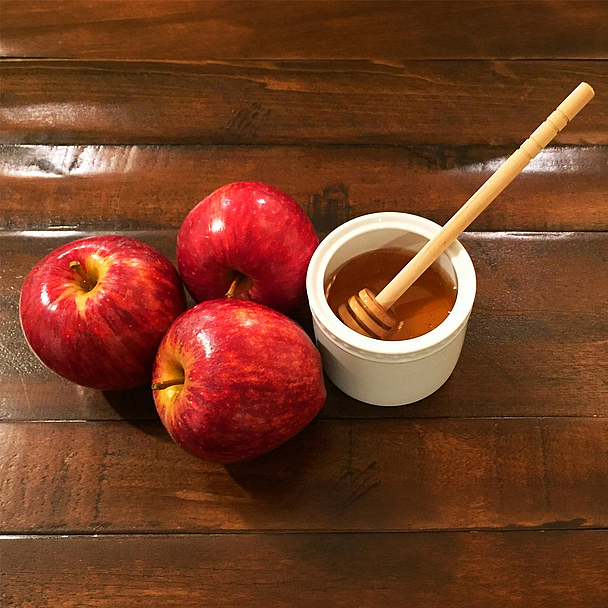 Drei rote Äpfel liegen neben einer Schale mit Honig und einem Honiglöffel auf einem dunklen Holztisch. Äpfel mit Honig gehören zu den traditionellen Speisen des jüdischen Neujahrsfestes Rosch Haschana.
