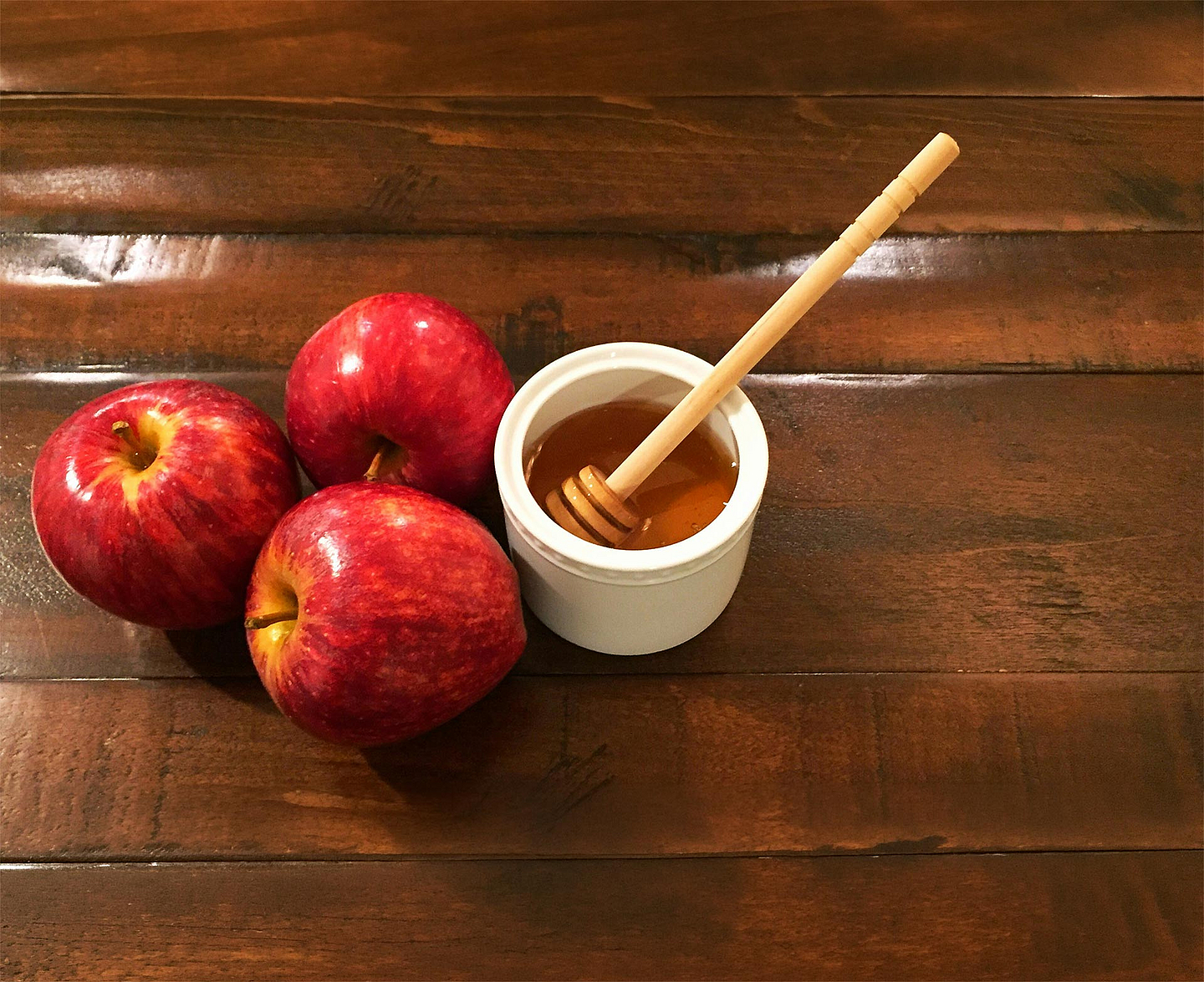 Äpfel und Honig stehen zu Rosch Haschana traditionell für die Hoffnung auf ein gutes und süßes neues Jahr. Drei rote Äpfel liegen neben einer Schale mit Honig und einem Honiglöffel auf einem dunklen Holztisch. Äpfel mit Honig gehören zu den traditionellen Speisen des jüdischen Neujahrsfestes Rosch Haschana.