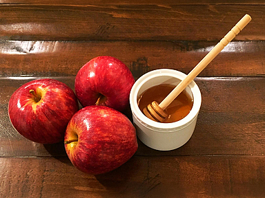 Drei rote Äpfel liegen neben einer Schale mit Honig und einem Honiglöffel auf einem dunklen Holztisch. Äpfel mit Honig gehören zu den traditionellen Speisen des jüdischen Neujahrsfestes Rosch Haschana.