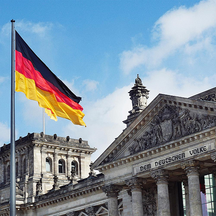 Das Bild zeigt die deutsche Nationalflagge in Schwarz-Rot-Gold, die an einem hohen Mast vor dem Reichstagsgebäude in Berlin weht. Rechts im Bild ist die Fassade des Reichstags zu sehen. Das Bild zeigt die deutsche Nationalflagge in Schwarz-Rot-Gold, die an einem hohen Mast vor dem Reichstagsgebäude in Berlin weht. Rechts im Bild ist die Fassade des Reichstags mit der Inschrift „Dem Deutschen Volke“ und klassizistischen Säulen sowie Reliefs erkennbar.