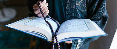 Unser Foto zeigt die heilige Schrift des Islams, den Koran. Zudem ist eine Misbaha zu sehen. Das ist eine Gebetskette, die den Beter oder die Beterin beim Sprechen bzw. dem Zählen von Gebetsformeln unterstützt. (Foto: Pexels/RODNAE)