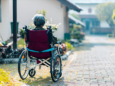 Eine Person sitzt in einem Rollstuhl und ist von hinten zu sehen. Der Rollstuhl steht auf einem gepflasterten Weg, der leicht nach vorne verläuft. Links und rechts des Weges sind Pflanzen und Sträucher zu erkennen. Im Hintergrund befinden sich Häuser mit hellen Fassaden und ein Dach mit grünen Ziegeln. Die Szene wirkt ruhig und zeigt eine Außenumgebung bei Tageslicht.