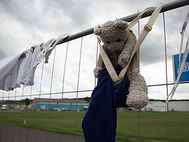 Ein Maschendrahtzaun mit aufgehängten Kleidungsstücken und einem Stofftier. Der Teddybär ist mit einem blauen Handtuch und einem beigen Band am Zaun befestigt. Im Hintergrund sind eine Wiese, einige Gebäude und ein bewölkter Himmel zu sehen.