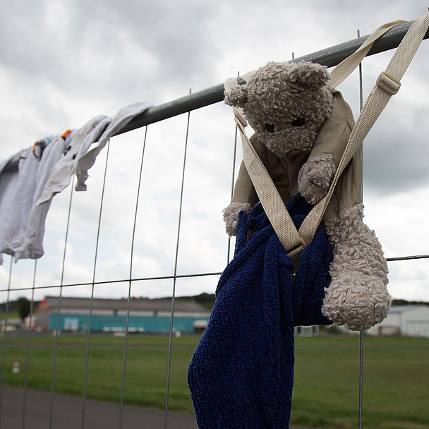 Ein Maschendrahtzaun mit aufgehängten Kleidungsstücken und einem Stofftier. Der Teddybär ist mit einem blauen Handtuch und einem beigen Band am Zaun befestigt. Im Hintergrund sind eine Wiese, einige Gebäude und ein bewölkter Himmel zu sehen.