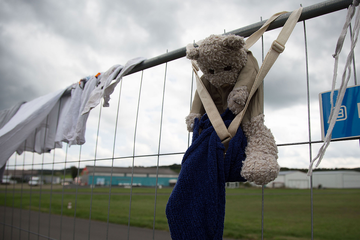 Ein Maschendrahtzaun mit aufgehängten Kleidungsstücken und einem Stofftier. Der Teddybär ist mit einem blauen Handtuch und einem beigen Band am Zaun befestigt. Im Hintergrund sind eine Wiese, einige Gebäude und ein bewölkter Himmel zu sehen.