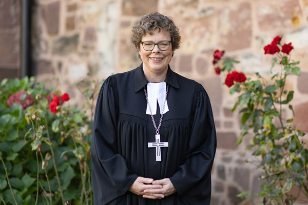 Dr. Beate Hofmann, Bischöfin der Evangelischen Kirche von Kurhessen-Waldeck (seit 2019) in schwarzer Amtstracht mit Kreuz vor historischer Steinwand und blühenden roten Rosen im Garten