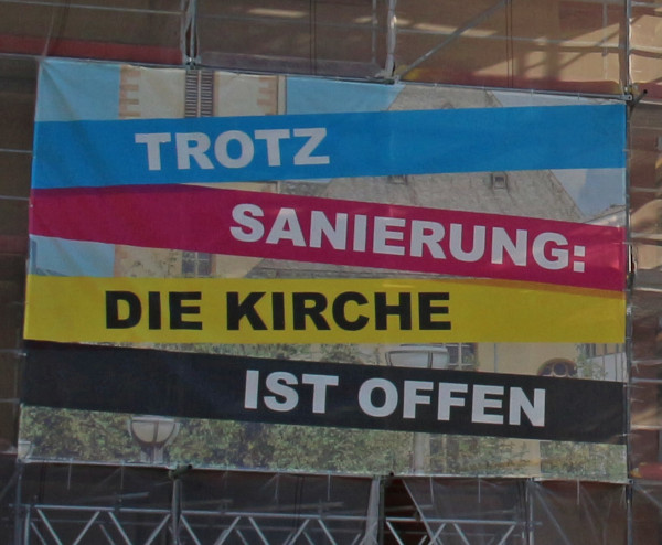 Werbeplane an einem Gerüst, das an der Fassade einer Kirche steht. Die Botschaft: Trotz Sanierung: Die Kirche ist offen.  