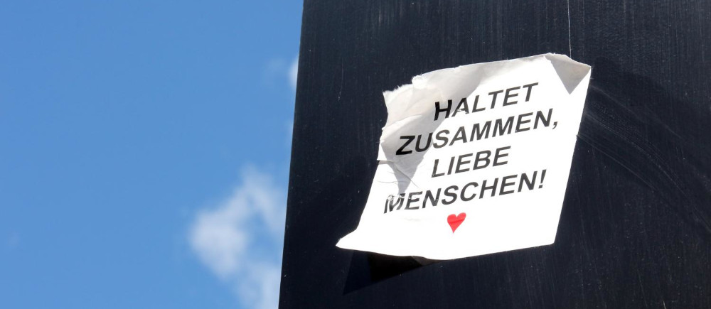 Zettel mit Aufschrift „Haltet zusammen, liebe Menschen!“ an schwarzer Wand, blauer Himmel. Foto: Peter Weidemann, pfarrbriefservice.de