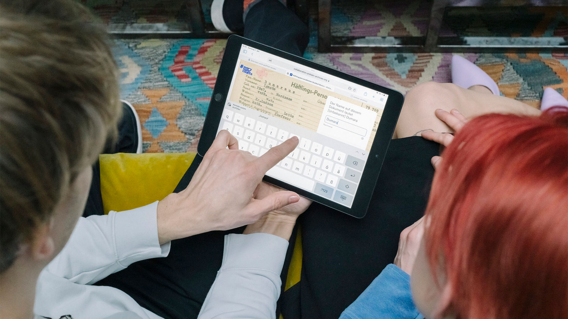 Das Bild zeigt einen Mann, der ein Tablet in der Hand hält und darauf zeigt. Darauf zu sehen ist eine Häftlingsakte. Neben ihm sitzt eine Frau. 