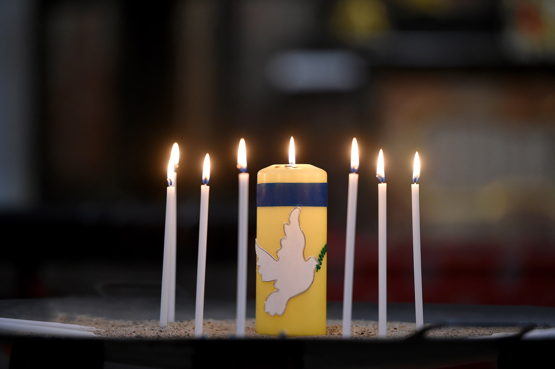 Das Bild zeigt Kerzen auf einem Altar. Darunter eine gelb-blaue Kerze mit einer Friedenstaube.