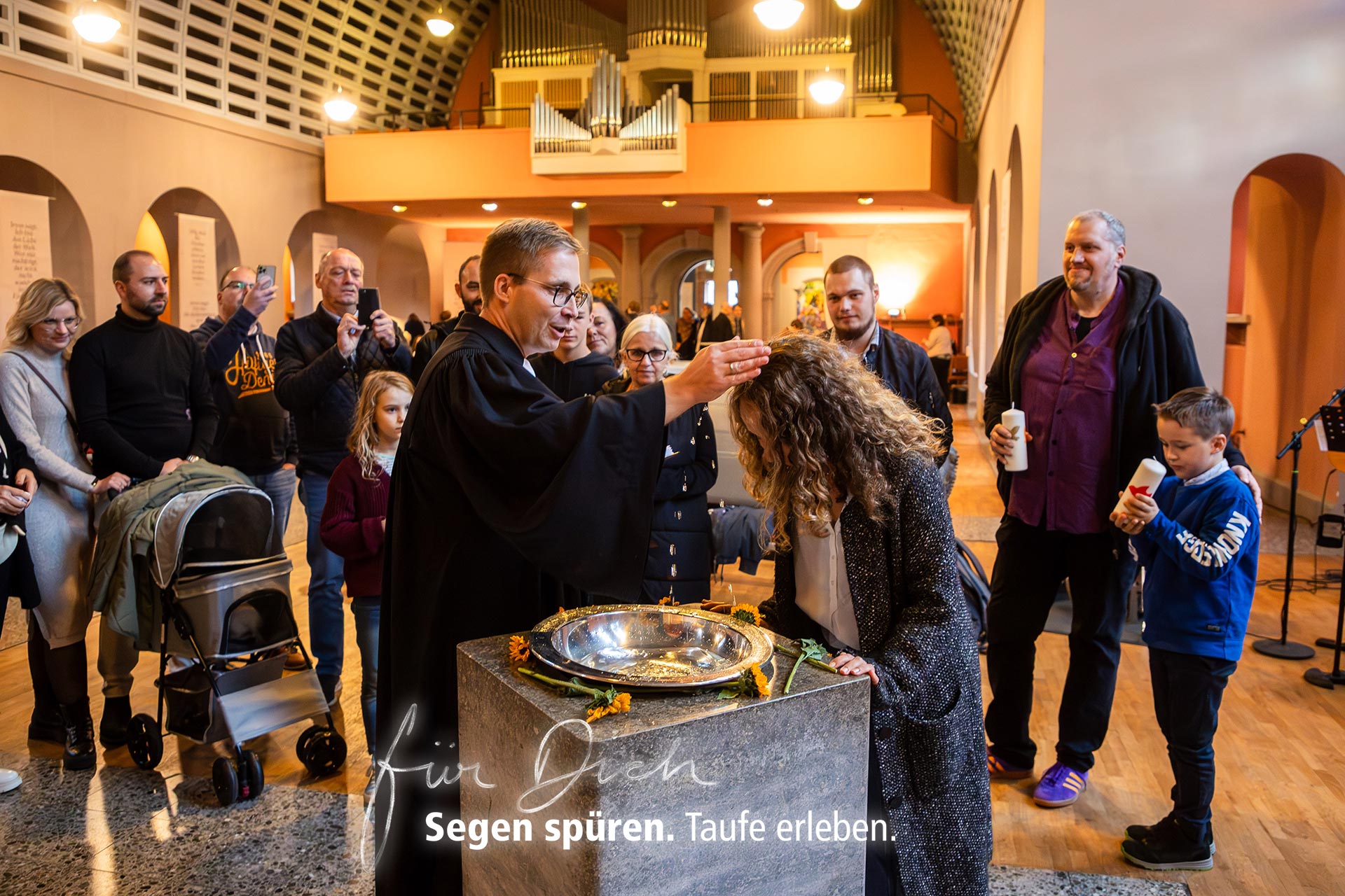 Eine Frau wird in einer evangelischen Kirche am Taufbecken von einem Pfarrer mit Wasser getauft. Um sie herum stehen Erwachsene und Kinder. Im Hintergrund sind Kirchenbänke, eine Orgel und ein Kinderwagen zu sehen. Text im Bild: ‚Für Dich. Segen spüren. Taufe erleben.