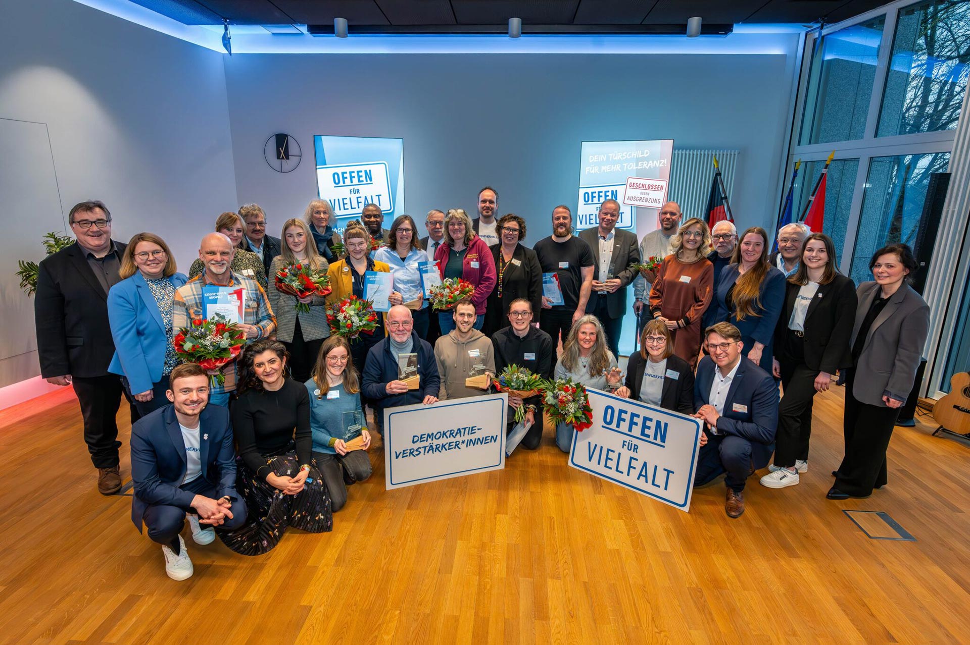 Das Bild zeigt eine Gruppe von etwa 30 Personen, die in einem Raum mit Holzboden und blauer Beleuchtung stehen. Einige Personen halten Blumensträuße und Schilder mit den Aufschriften „Offen für Vielfalt“ und „Demokratie-Verstärker*innen“. Im Hintergrund sind weitere Schilder zu sehen. In ganz links steht Kassels Regierungspräsident Mark Weinmeister, in der ersten Reihe rechts Christoph Lübcke, Sohn des ermordeten Kasseler Regierungspräsidenten Walter Lübcke.