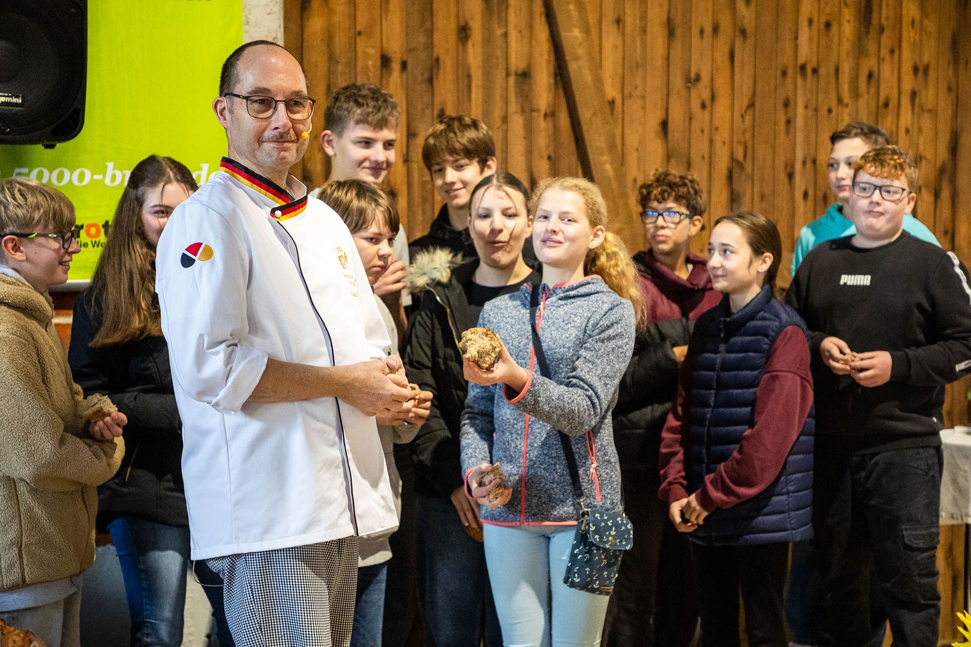 Bäckermeister Bernd Riede steht in einer Scheune und hält ein Brot. Um ihn herum stehen Konfirmandinnen und Konfirmanden aus Vellmar, die ebenfalls Brote in den Händen halten.